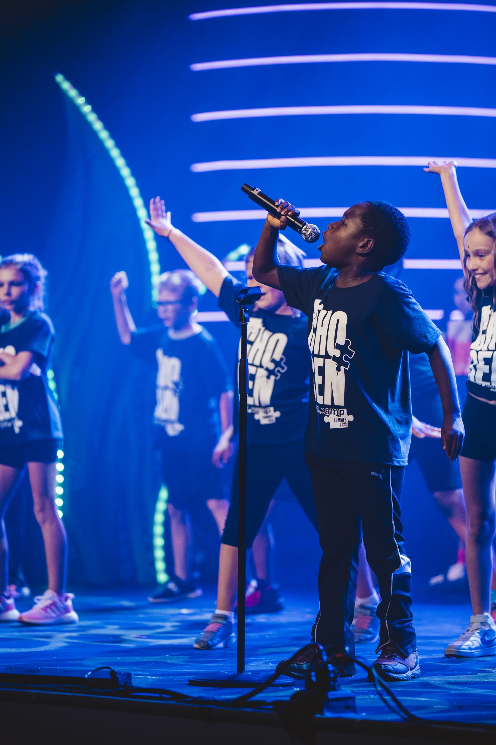 A young performer in a Chosen tee sings into a microphone under bright blue and green stage lights