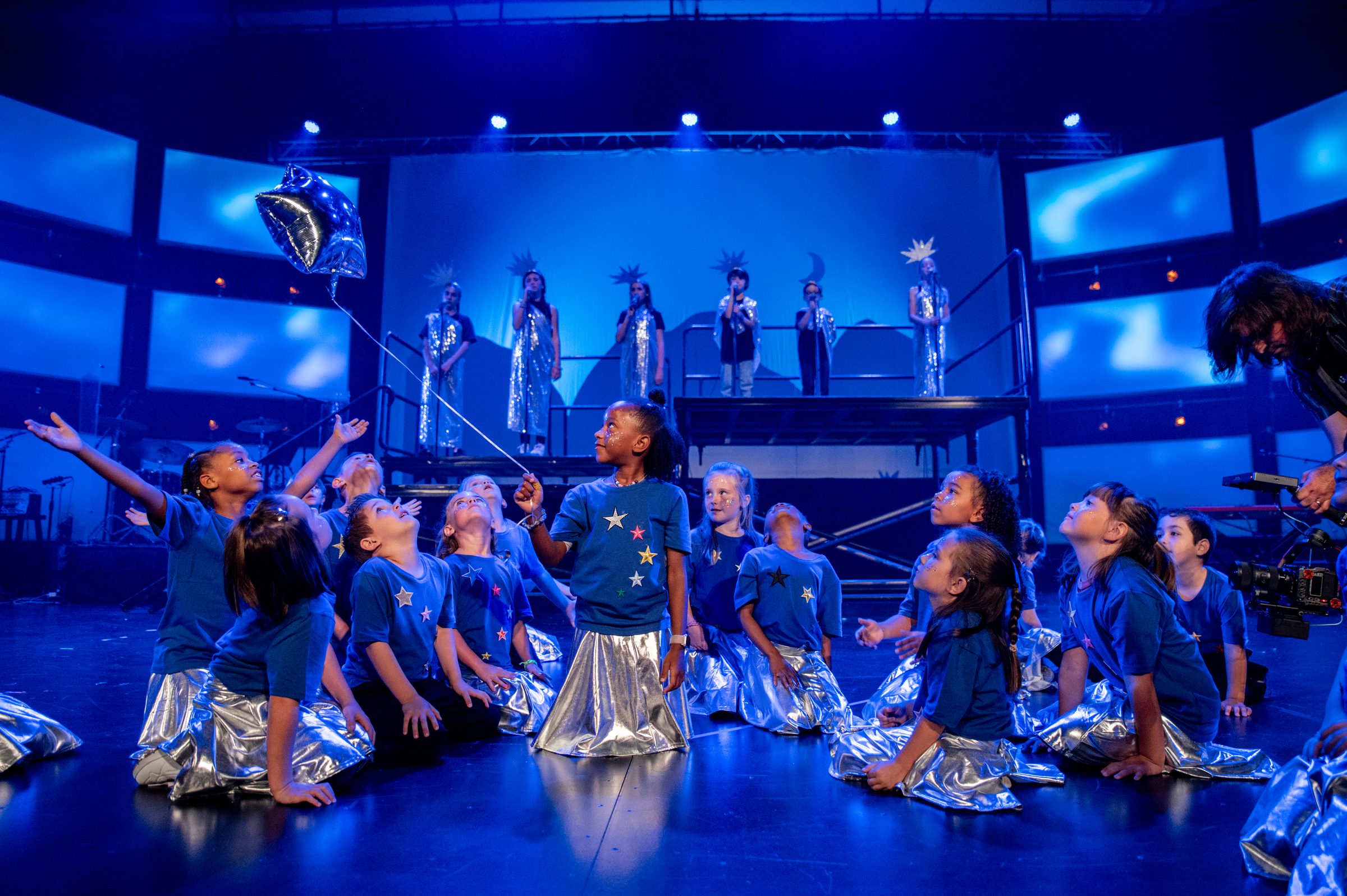 A student holds a silver star-shaped balloon amid a cast of young performers in starry costumes on a blue-lit stage