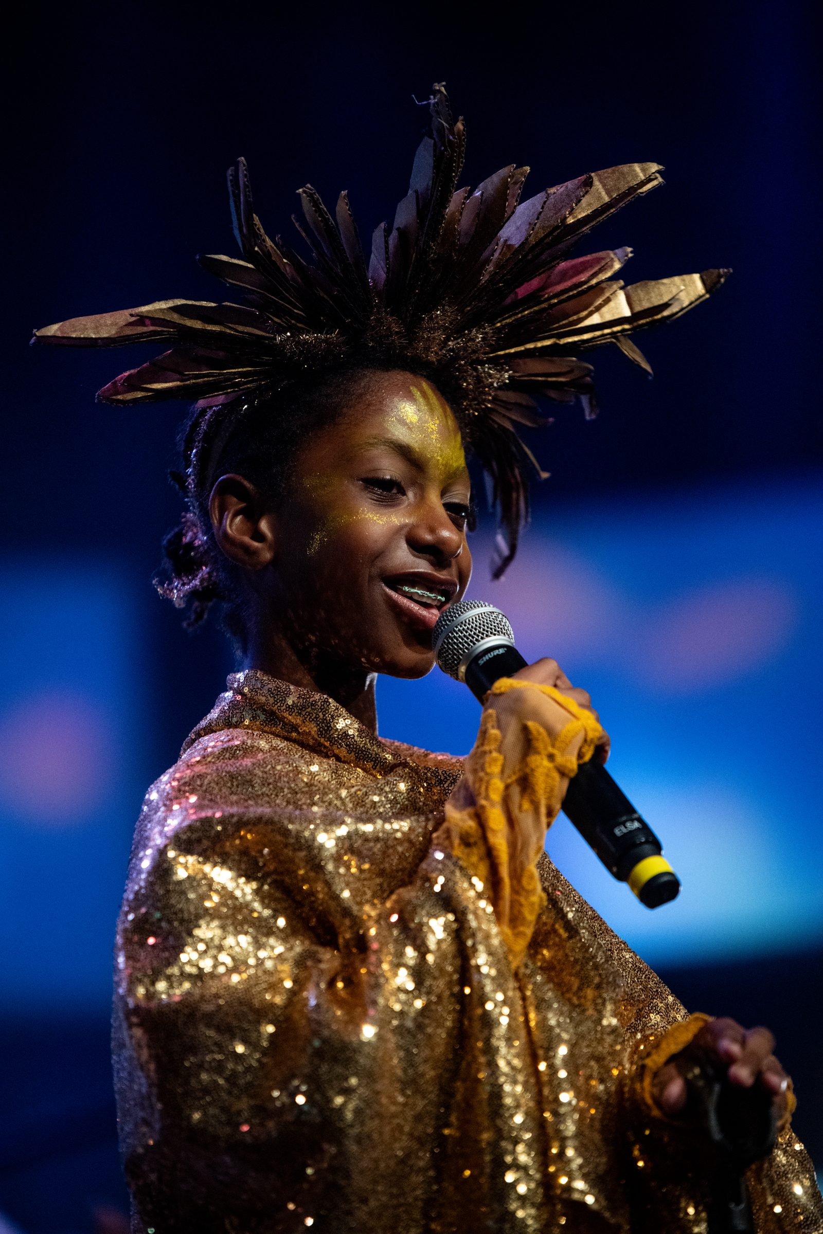 A young singer in a gold sequined costume performs into a microphone under stage lights