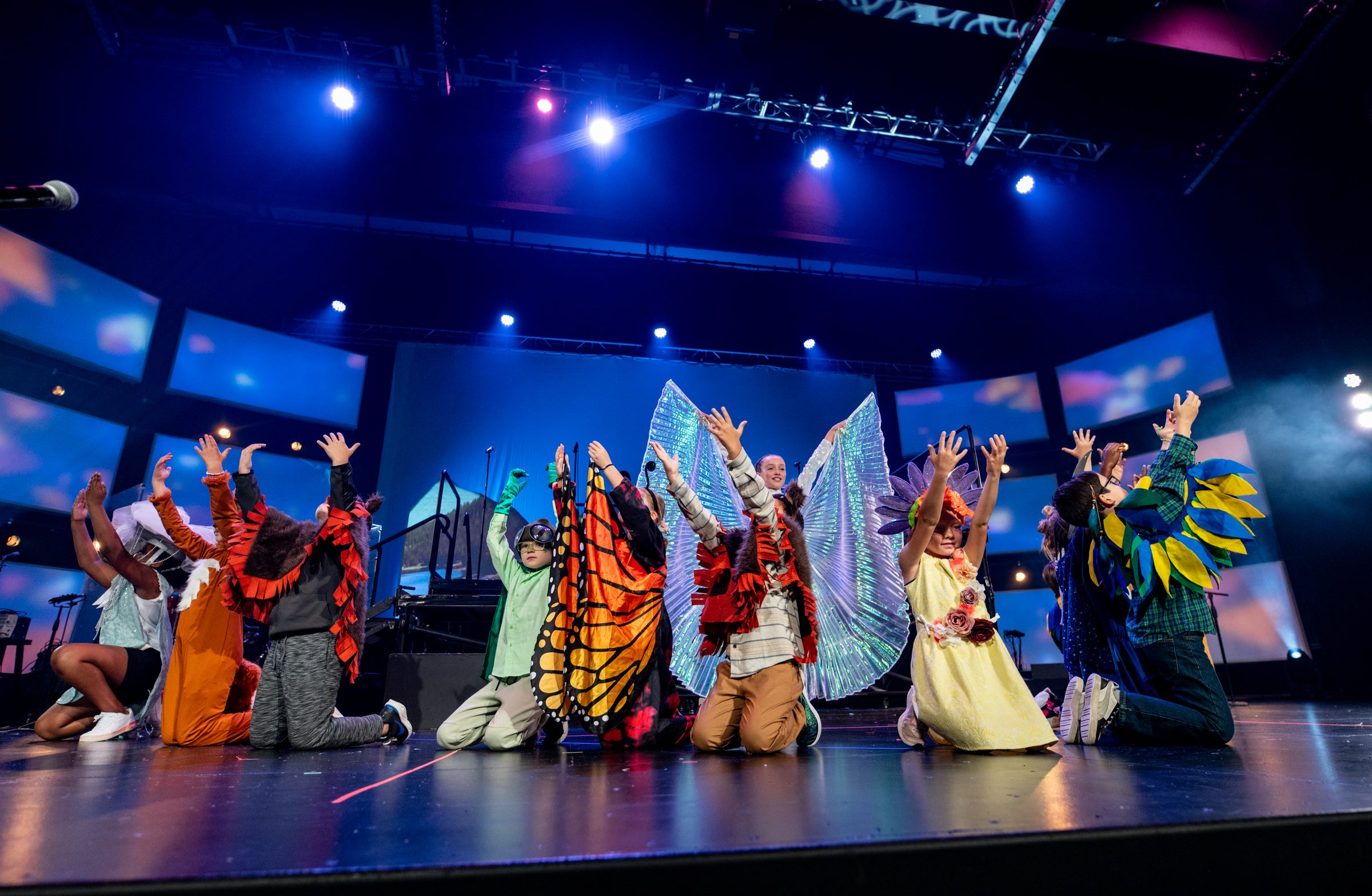 A row of young performers in butterfly, sunflower, and bird costumes raise their arms mid-performance