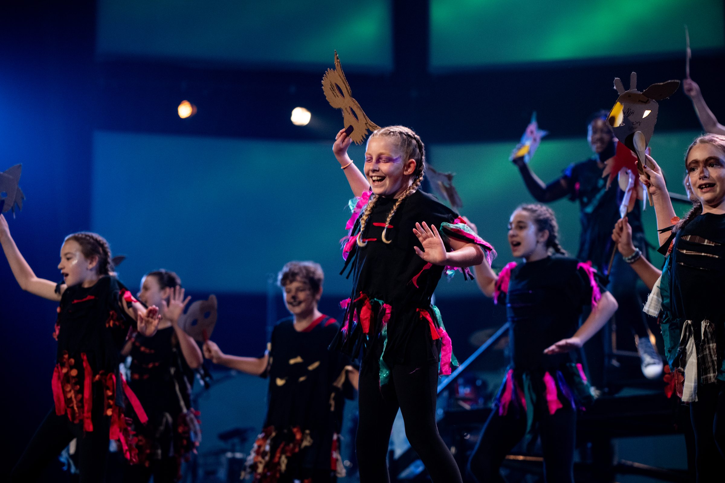 A young performer laughs mid-dance holding a handmade paper mask aloft