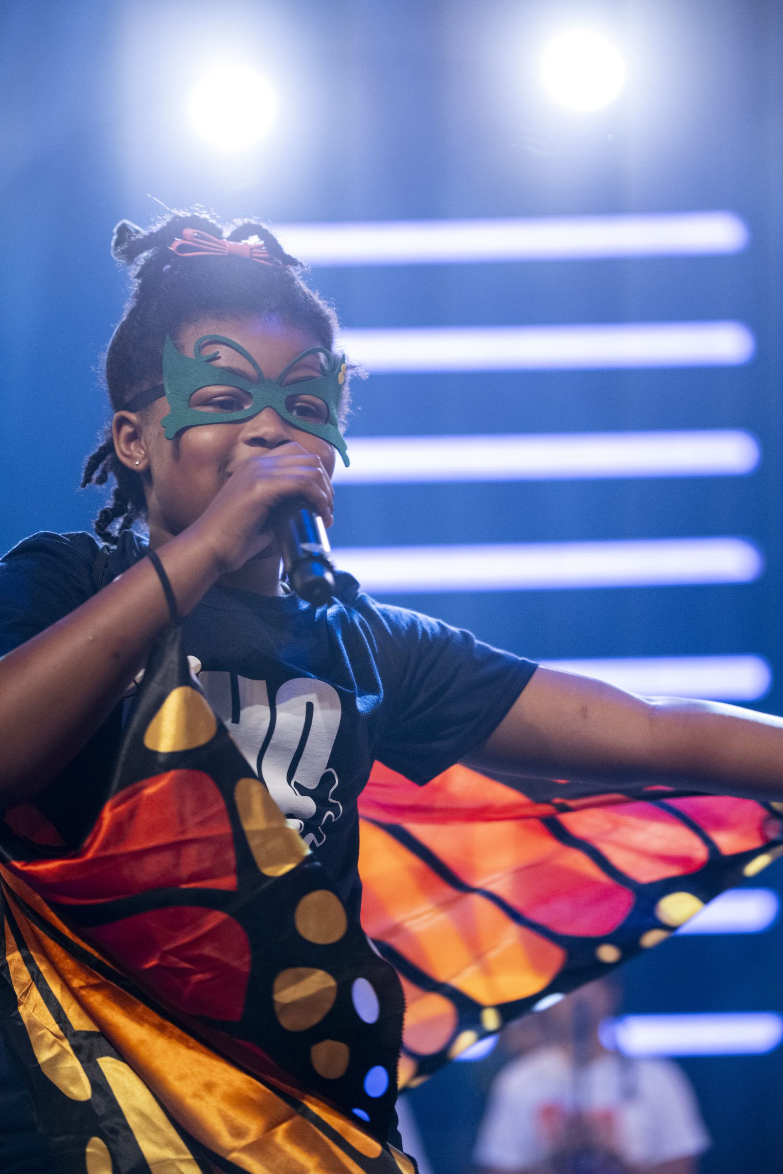 A young Summer Camp performer in a butterfly cape and green mask sings into a microphone, wings spread wide