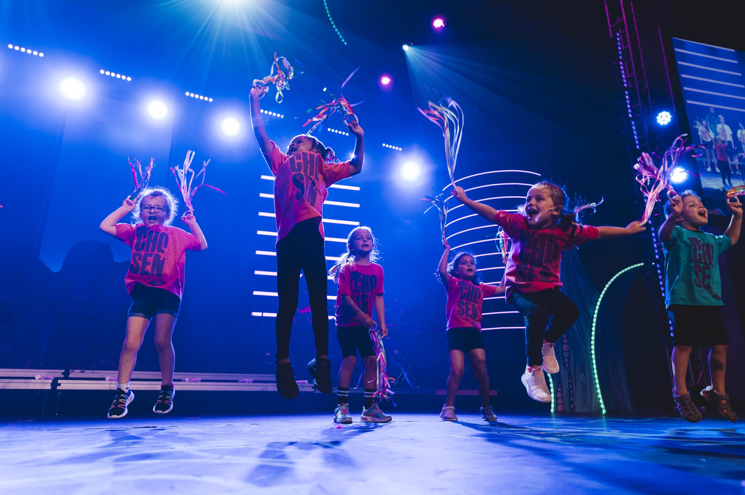 Young children in pink and green Chosen t-shirts jump joyfully with ribbon streamers on a magenta-lit stage