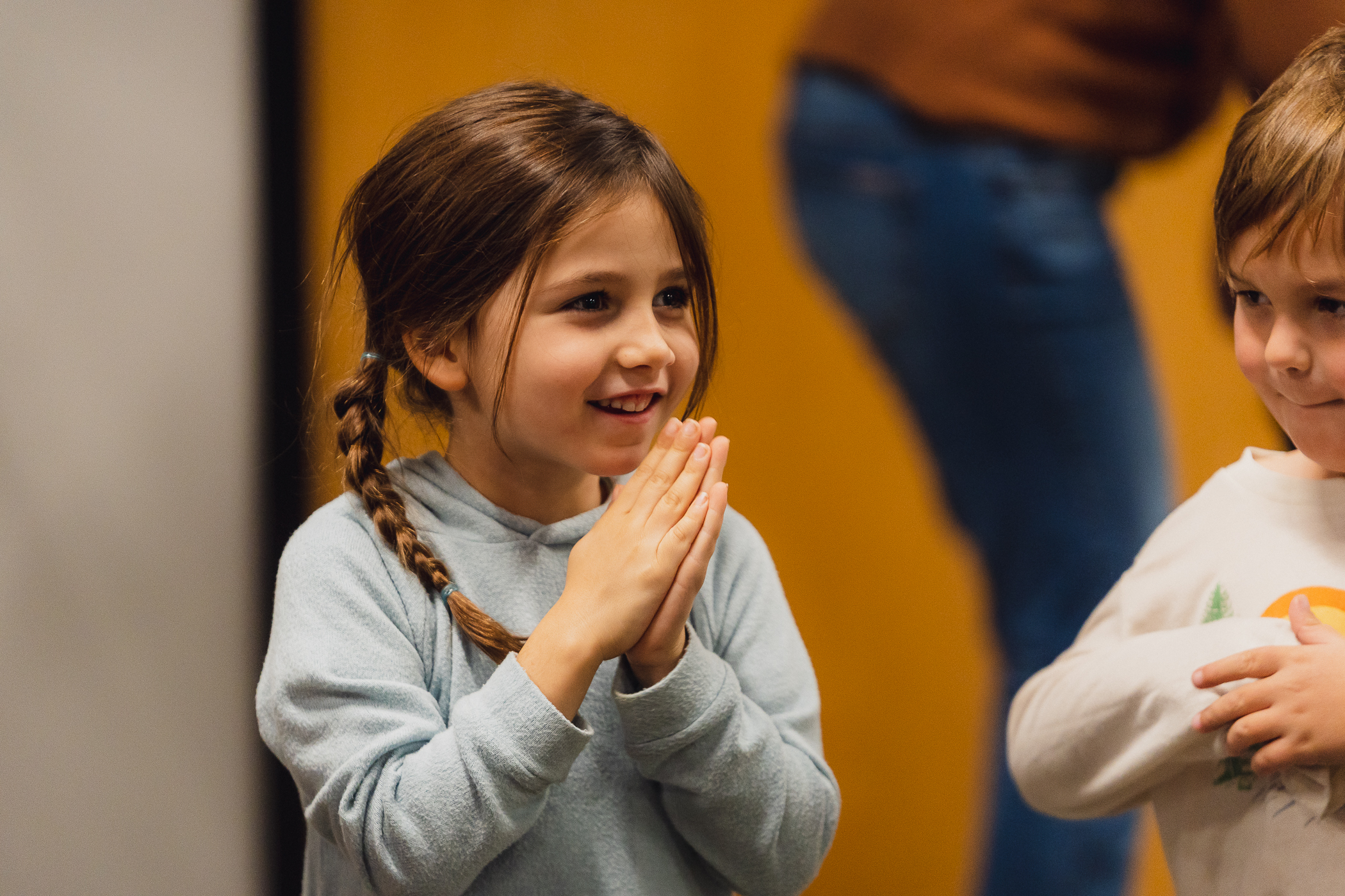 A young girl in a light blue sweatshirt smiles and claps her hands together in class