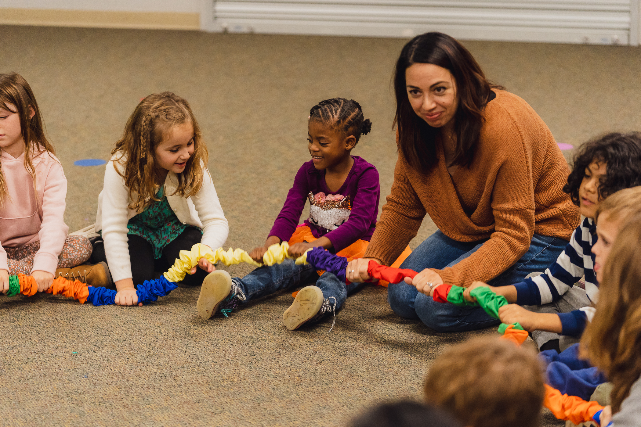A teacher and a circle of young children play with a rainbow stretchy band on the classroom floor