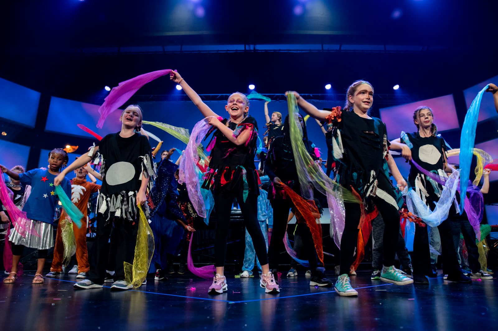 A line of Kaleo Kids performers dance across the stage waving colorful ribbons and scarves under blue stage lights