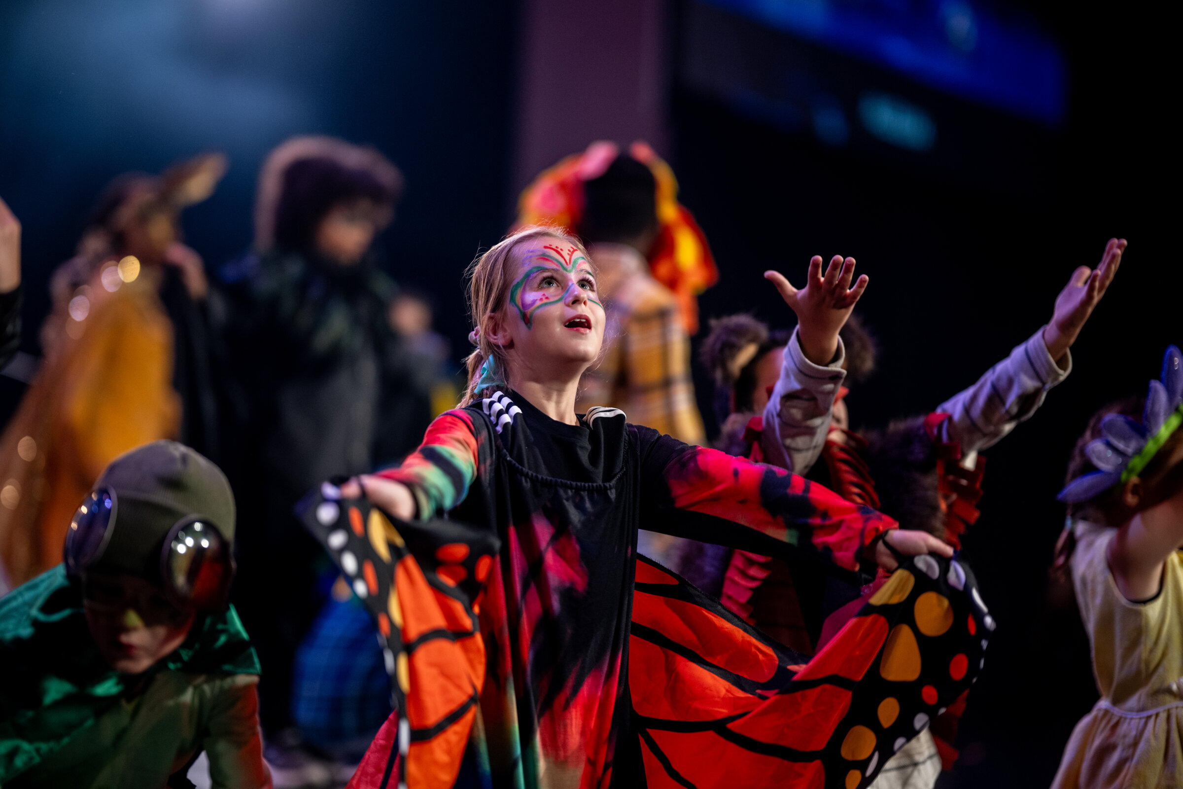 A young performer in butterfly wings, arms outstretched mid-performance under warm stage light