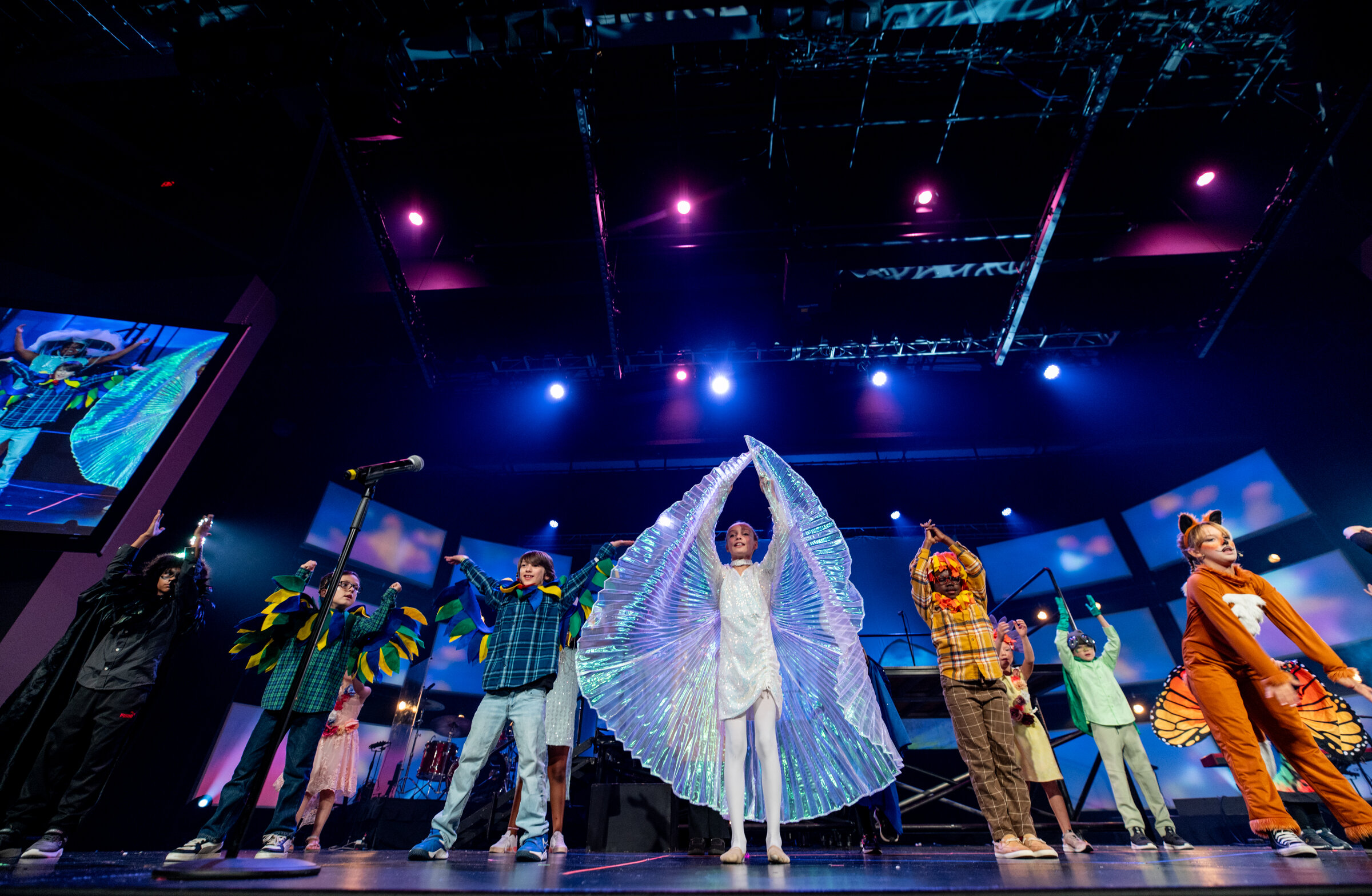 A wide low-angle stage shot with the full ensemble arms raised, a dancer in luminous wings at center under the lighting rig