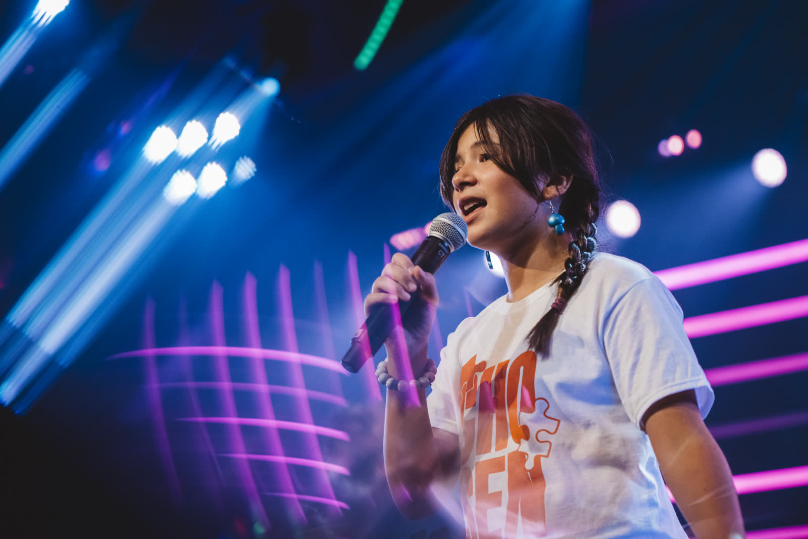 A young Kaleo Kids performer sings into a microphone on stage, framed by streaks of magenta and blue concert lighting
