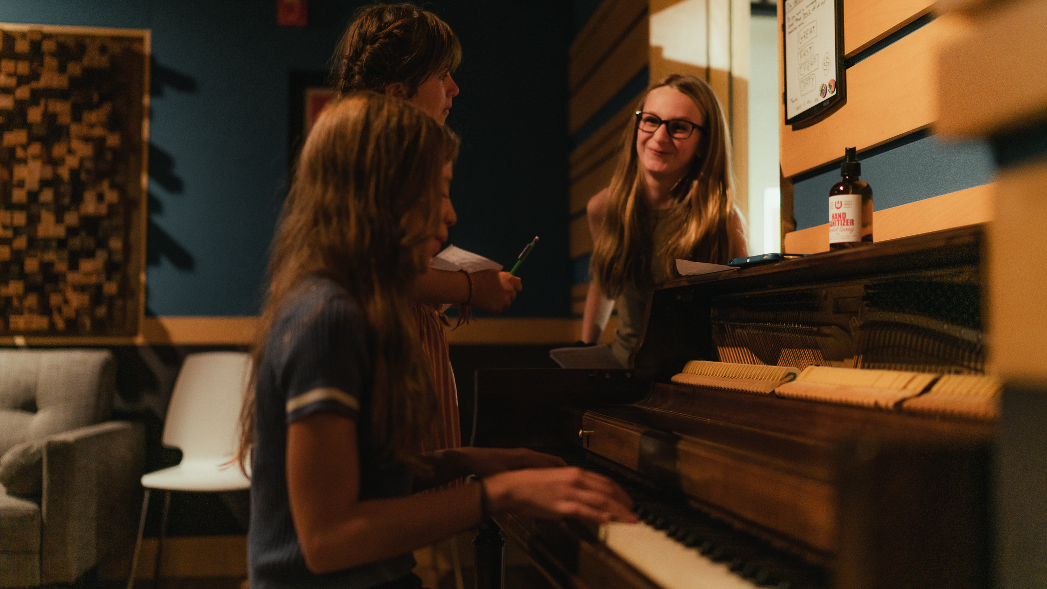 A young songwriter smiles from behind the upright piano while a friend plays through an idea